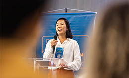 A young Asian woman holding a microphone is on stage giving a presentation in front of a blue background. In the foreground are blurry images of audience members listening