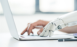 A human hand and white robot hand type side-by-side on a laptop keyboard on a white table top, with a black pen sitting to the side and soft white light coming through windows in soft focus in the background