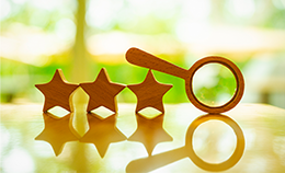 Three wooden stars sit on a reflective surface, with a wooden magnifying glass propped on the star at right by its handle. Their shapes are reflected beneath them, with softly blurred green and yellow shapes visible through a window in the background