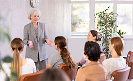 An older, silver-haired woman in a gray business suit stands before a class of college students, sharing her story
