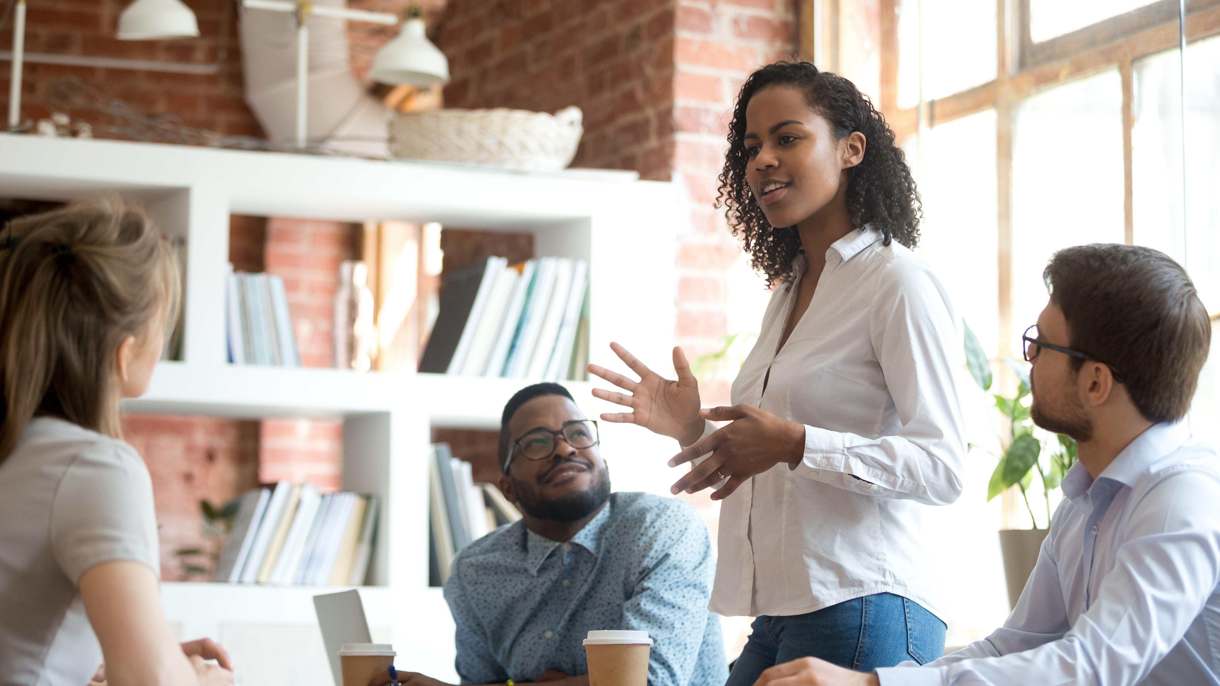 Diverse group of people in business setting gathered around a table with a laptop, listen while standing Black woman teaches.  