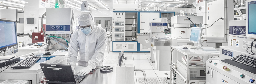 A researcher in hooded white cleanroom coveralls, mask and gloves types on a laptop in a white-tiled cleanroom packed with high-tech equipment.