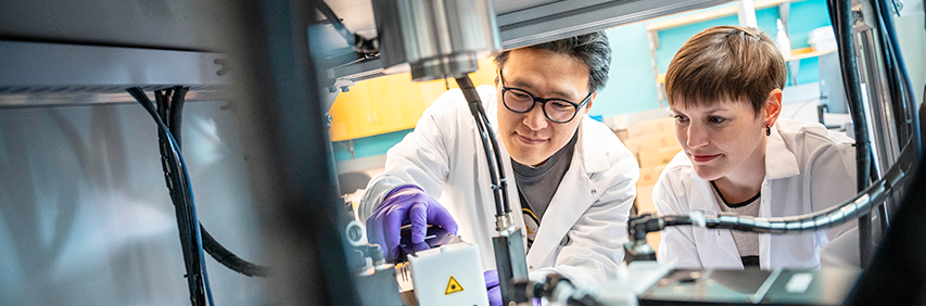 Two scientists in lab coats and gloves examine the contents of a metal chamber equipped with high-tech components.