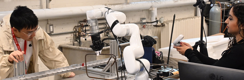 Un bras robotisé à plusieurs articulations tient un rectangle formé à partir d’une barre d’armature. À droite, une jeune femme tient une petite boîte blanche. À gauche, un jeune homme observe le support dans lequel la barre d’armature est insérée.