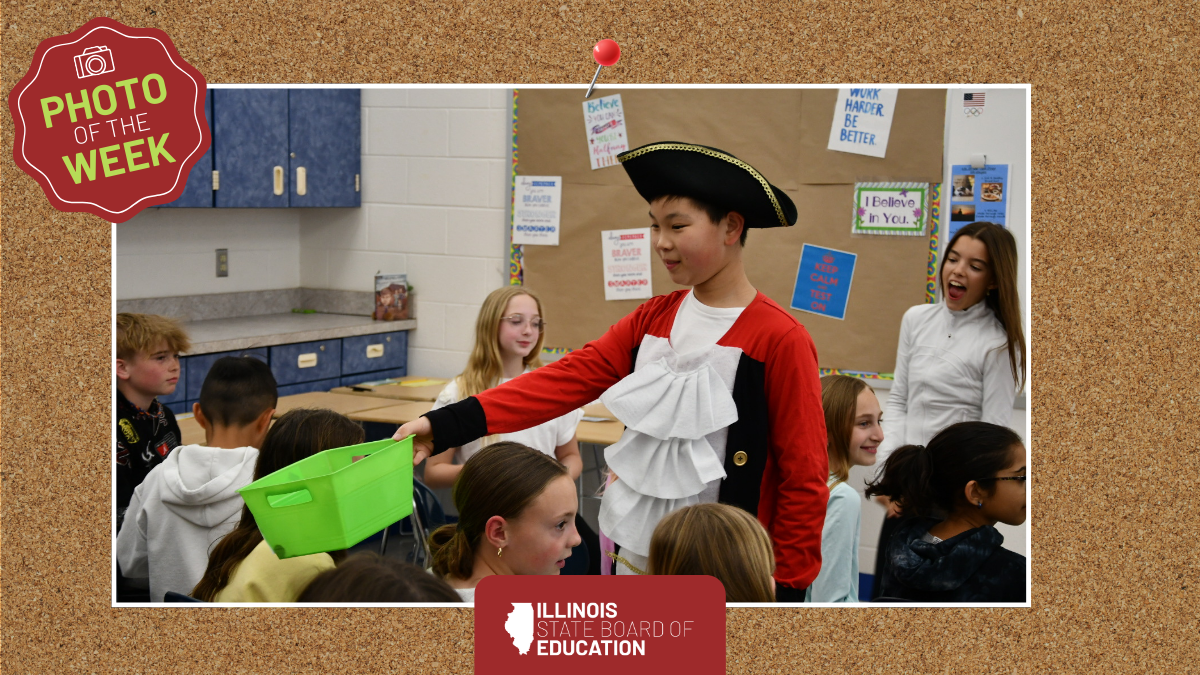 An elementary student wearing a red colonial soldier costume holds out a green bin to classmates.