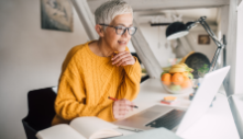 person wearing glasses sitting at a desk with a laptop and holding a pen